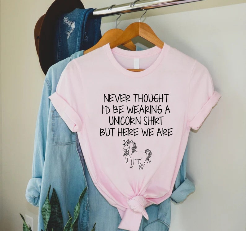 May include: Pink t-shirt with a black unicorn graphic and the text "Never thought I'd be wearing a unicorn shirt but here we are."