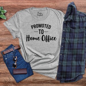 May include: A grey t-shirt with the text "PROMOTED TO Home Office" in black, alongside blue jeans, a brown belt, sunglasses, and a wallet. A blue and green plaid shirt is also included. A small succulent plant is in the top left corner.