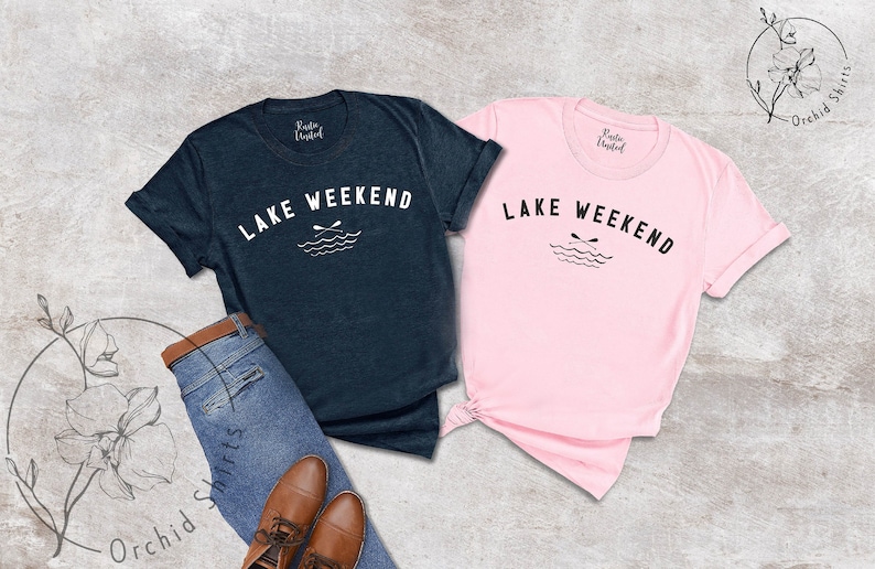 以下が含まれることがあります： 紺色とピンク色の2枚のTシャツ。白で「LAKE WEEKEND」の文字がプリントされています。テキストの下には、波と交差したオールのグラフィックがあります。Tシャツはジーンズと茶色の靴と一緒に表示されています。