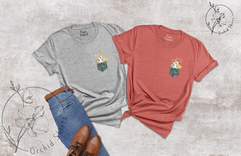 May include: Two t-shirts, one gray and one coral, with a pocket design featuring a unicorn and the text "Sunshine's in my pocket." The shirts are displayed with blue jeans and brown shoes, suggesting casual wear.