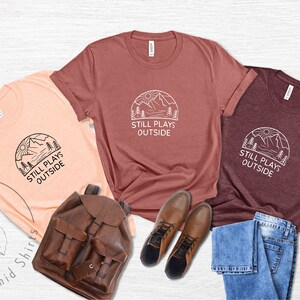 May include: Three t-shirts with a mountain scene and the text "Still Plays Outside". The shirts are in peach, maroon, and dark grey. The shirts are laid out on a white surface with a brown leather backpack, blue jeans, and brown boots.