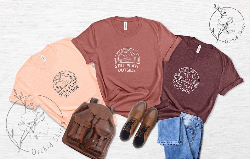 May include: Three t-shirts with a mountain scene and the text "Still Plays Outside". The shirts are in peach, maroon, and dark grey. The shirts are laid out on a white surface with a brown leather backpack, blue jeans, and brown boots.