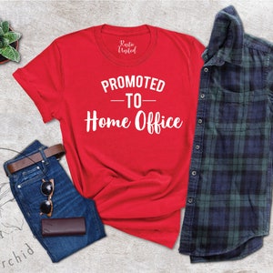 May include: A red t-shirt with the text "PROMOTED - TO - Home Office" in white, alongside blue jeans, a brown belt, sunglasses, and a wallet. A green and navy plaid shirt is also included.