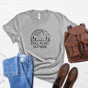 May include: A heather grey t-shirt with a black graphic of a mountain range, sun, and trees. The text "Still Plays Outside" is printed below the graphic.