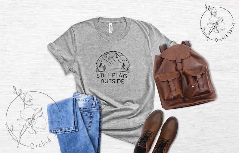 May include: A heather grey t-shirt with a black graphic of a mountain range, sun, and trees. The text "Still Plays Outside" is printed below the graphic.
