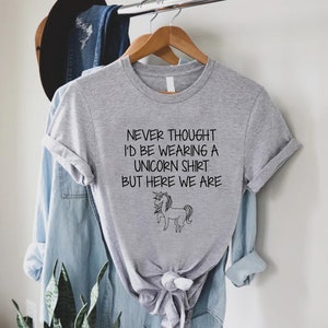 May include: Gray t-shirt with a black and white unicorn illustration and the text "Never thought I'd be wearing a unicorn shirt but here we are."