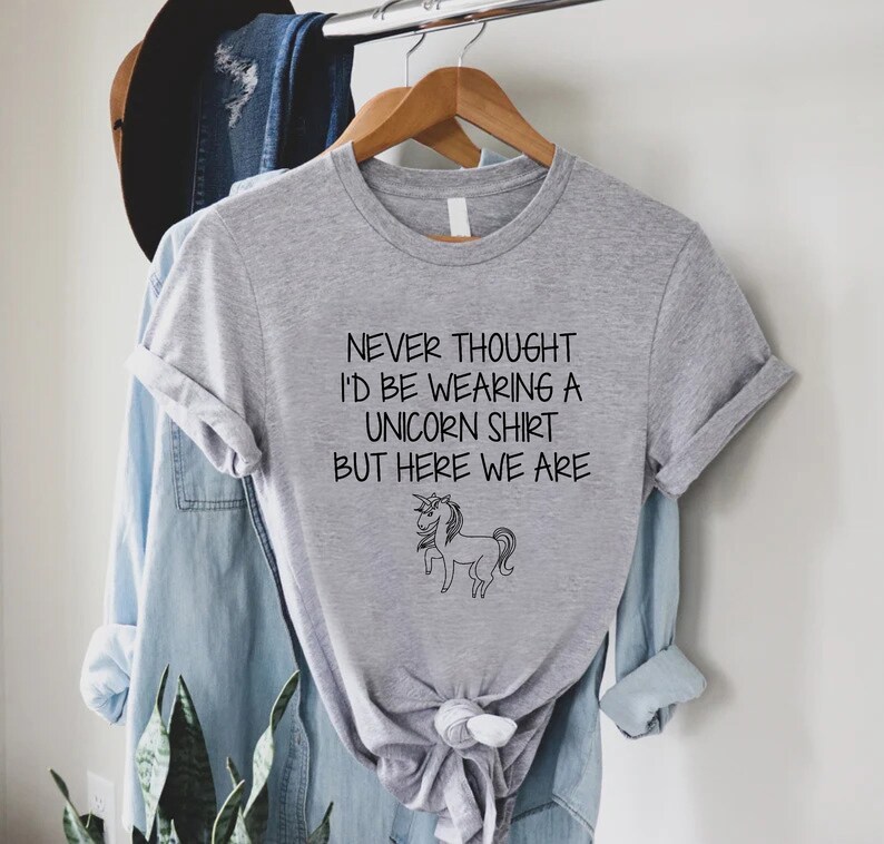 May include: Gray t-shirt with a black and white unicorn illustration and the text "Never thought I'd be wearing a unicorn shirt but here we are."