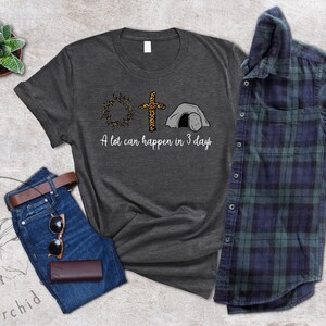 K&ouml;nnte beinhalten: Ein dunkelgraues T-Shirt mit einem Motiv aus einer Dornenkrone, einem Kreuz mit Leopardenmuster und einem Grab. Der Text "A lot can happen in 3 days" ist unter dem Motiv gedruckt.