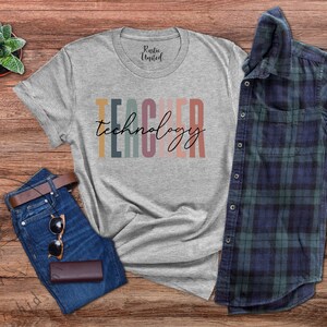 Pu&ograve; includere: Una maglietta grigia con il testo "TEACHER technology" in strisce verticali colorate. La maglietta &egrave; appoggiata su una superficie di legno con un paio di jeans, occhiali da sole e una camicia a quadri.