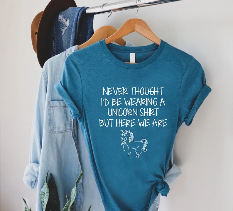 May include: Teal blue t-shirt with white text that reads "Never thought I'd be wearing a unicorn shirt but here we are" and a white outline drawing of a unicorn.