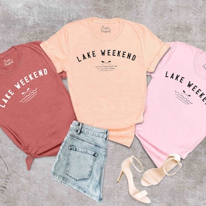 以下が含まれることがあります： コーラル、ピーチ、ピンクの半袖Tシャツ3枚。それぞれに「LAKE WEEKEND」の文字と、オールと波のグラフィックがプリントされています。シャツはデニムショーツとベージュのハイヒールと組み合わせています。