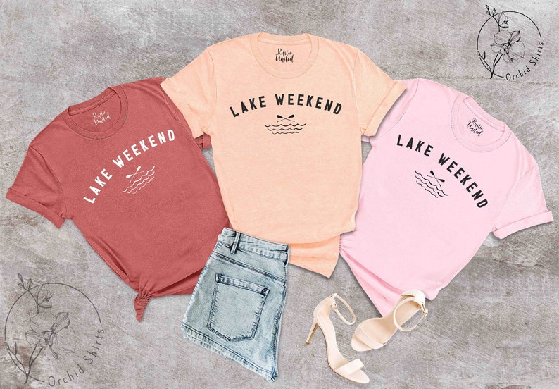 以下が含まれることがあります： コーラル、ピーチ、ピンクの半袖Tシャツ3枚。それぞれに「LAKE WEEKEND」の文字と、オールと波のグラフィックがプリントされています。シャツはデニムショーツとベージュのハイヒールと組み合わせています。