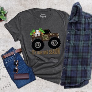 May include: Dark grey t-shirt featuring a monster truck with a camouflage design, carrying a bunny and carrots. The text "IT'S HUNTING SEASON!" is below the truck. The shirt is paired with blue jeans, a brown belt, sunglasses, and a wallet.
