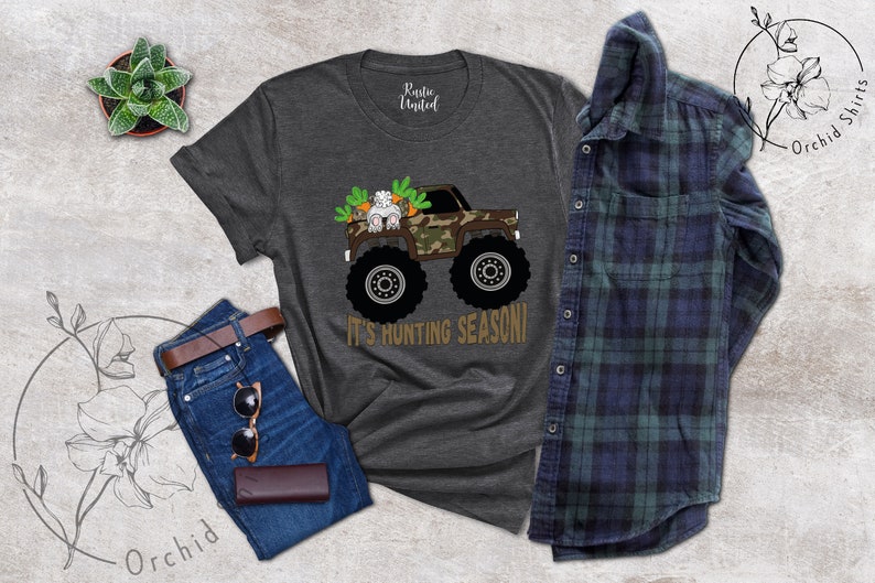 May include: Dark grey t-shirt featuring a monster truck with a camouflage design, carrying a bunny and carrots. The text "IT'S HUNTING SEASON!" is below the truck. The shirt is paired with blue jeans, a brown belt, sunglasses, and a wallet.
