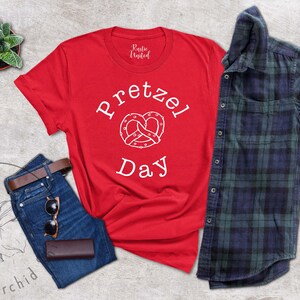 Op de afbeelding: Rood T-shirt met witte tekst die "Pretzel Day" zegt en een witte omtrek van een pretzel.