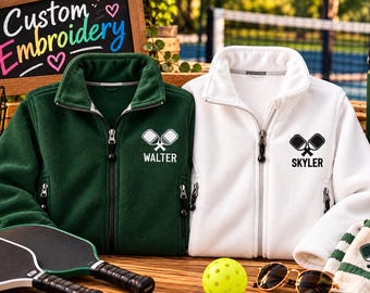 May include: A dark green fleece jacket with the name WALTER and a pickleball paddle graphic, and a white fleece jacket with the name SKYLER and a pickleball paddle graphic. Both jackets feature full-length zips and side pockets.