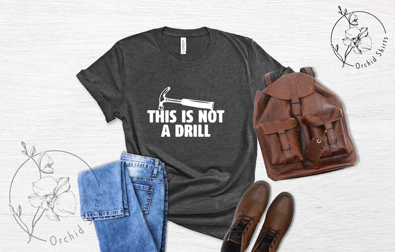 May include: A dark gray t-shirt with a white hammer and the text "This is not a drill" printed on the front. The shirt is displayed on a white wooden background with a pair of blue jeans, a brown leather backpack, and brown leather boots.