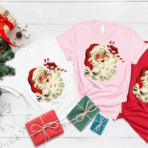 May include: Three t-shirts with a vintage illustration of Santa Claus holding a candy cane. The shirts are white, pink, and red.