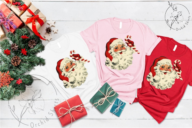 May include: Three t-shirts with a vintage illustration of Santa Claus holding a candy cane. The shirts are white, pink, and red.