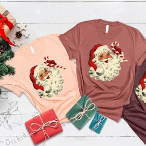 May include: Three t-shirts with a vintage illustration of Santa Claus holding a candy cane. The shirts are in peach, maroon, and burgundy.