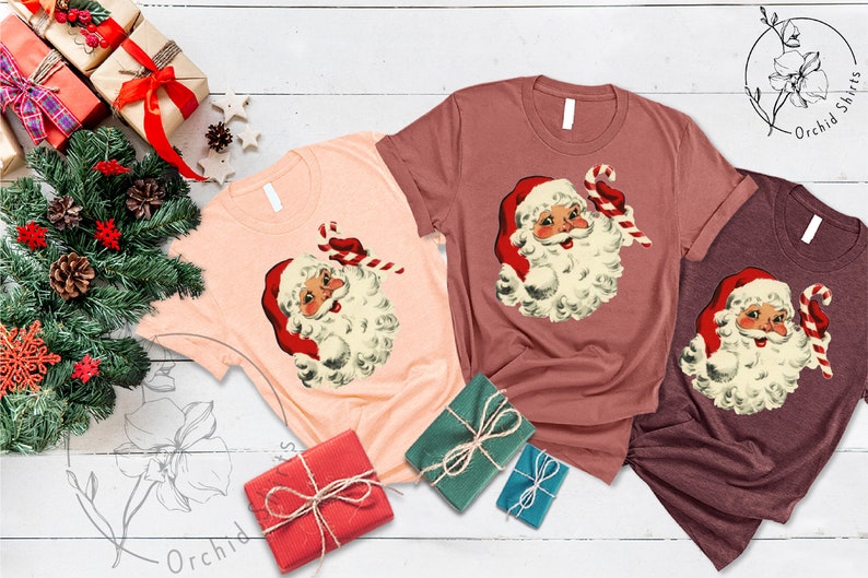 May include: Three t-shirts with a vintage illustration of Santa Claus holding a candy cane. The shirts are in peach, maroon, and burgundy.