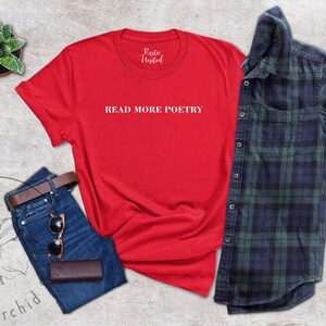 Op de afbeelding: Rood T-shirt met de tekst "READ MORE POETRY" in witte letters. Het shirt ligt op een lichtgrijze ondergrond met een blauwe spijkerbroek, een bruine leren portemonnee, een zonnebril en een klein potplantje.