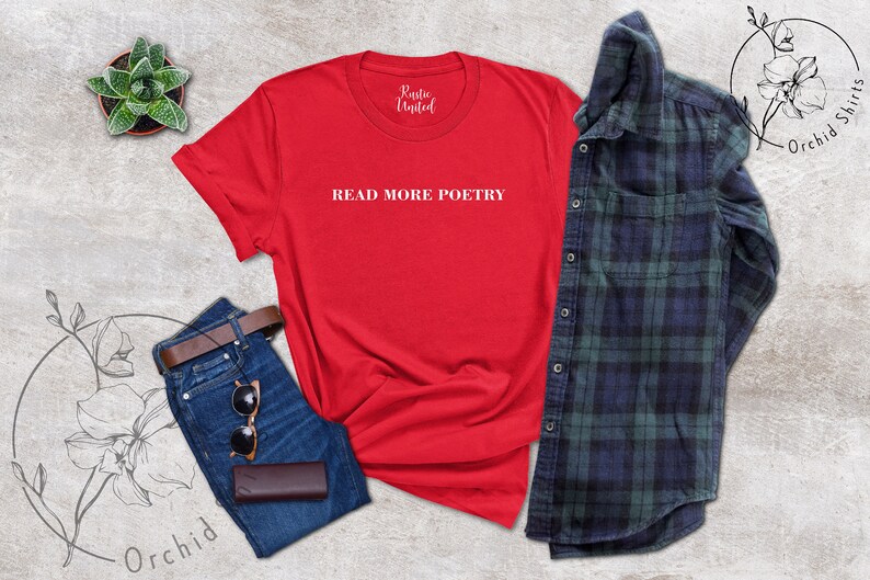 Op de afbeelding: Rood T-shirt met de tekst "READ MORE POETRY" in witte letters. Het shirt ligt op een lichtgrijze ondergrond met een blauwe spijkerbroek, een bruine leren portemonnee, een zonnebril en een klein potplantje.