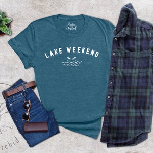 以下が含まれることがあります： 「LAKE WEEKEND」の白文字とオールと波のグラフィックが入った、ティール色のTシャツ。青いジーンズ、茶色のベルト、サングラス、茶色の財布と組み合わせています。チェック柄のシャツも含まれています。