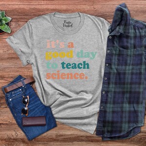 Op de afbeelding: Een grijs T-shirt met de tekst "it's a good day to teach science." in kleurrijke letters. Het shirt ligt op een houten oppervlak met een spijkerbroek, een zonnebril en een geruit overhemd.