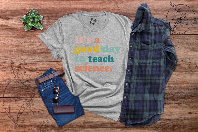 Op de afbeelding: Een grijs T-shirt met de tekst "it's a good day to teach science." in kleurrijke letters. Het shirt ligt op een houten oppervlak met een spijkerbroek, een zonnebril en een geruit overhemd.