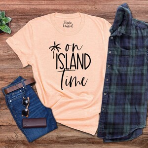 May include: A peach-colored t-shirt with black text that reads "*on island time". The shirt is laid out on a wooden surface with a pair of blue jeans, a brown leather belt, sunglasses, and a plaid shirt.