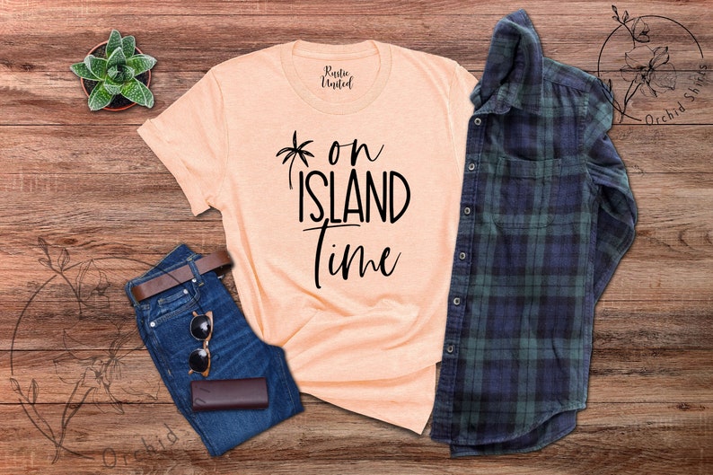 May include: A peach-colored t-shirt with black text that reads "*on island time". The shirt is laid out on a wooden surface with a pair of blue jeans, a brown leather belt, sunglasses, and a plaid shirt.