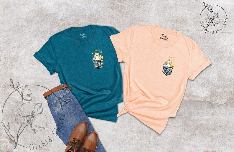May include: Two t-shirts, one teal and one peach, each with a pocket featuring a unicorn design. The shirts are displayed with blue jeans and brown leather shoes, suggesting a casual style. The text "Teacher's Pet" is above the unicorn.