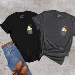 May include: Two t-shirts, one black V-neck and one charcoal crew neck, each with a pocket design featuring a unicorn and the text "Teacher's Pet." The shirts are displayed with blue jeans and brown shoes.