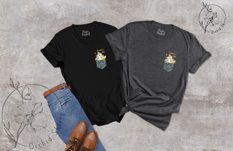 May include: Two t-shirts, one black V-neck and one charcoal crew neck, each with a pocket design featuring a unicorn and the text "Teacher's Pet." The shirts are displayed with blue jeans and brown shoes.