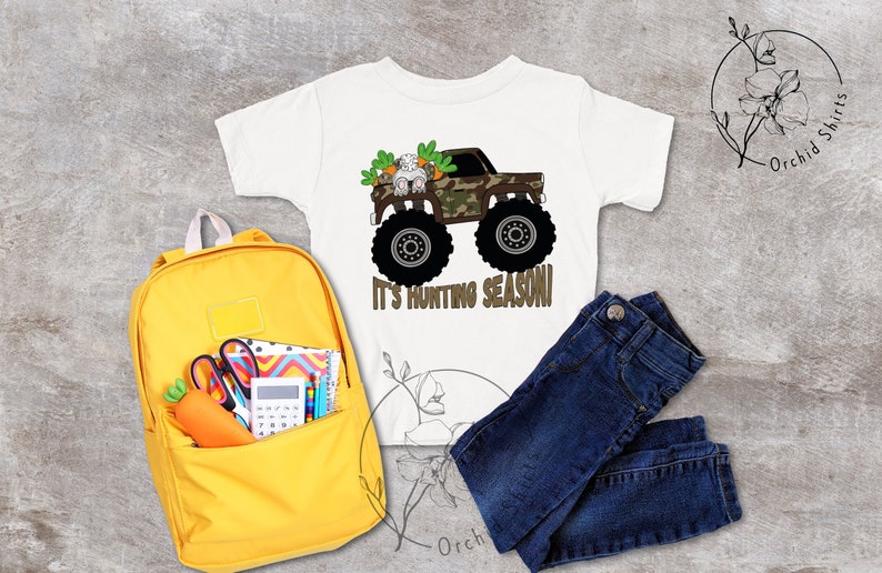 May include: White t-shirt with a monster truck graphic, camouflage cab, and the text "It's Hunting Season!". Includes a yellow backpack, blue jeans, and school supplies. The shirt features a cartoon bunny and carrots.
