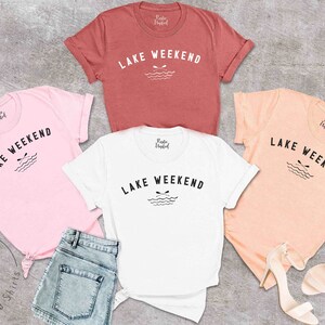 以下が含まれることがあります： ピンク、白、ピーチ、ラストカラーの半袖Tシャツ5枚。各シャツには、「LAKE WEEKEND」の文字と、オールと波のグラフィックが描かれています。シャツは、デニムショーツとサンダルと一緒に表示されています。