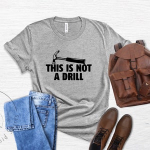 May include: A gray t-shirt with a black hammer and the text "This is not a drill". The shirt is laid out on a wooden surface with a pair of jeans, a brown leather backpack, and brown leather boots.