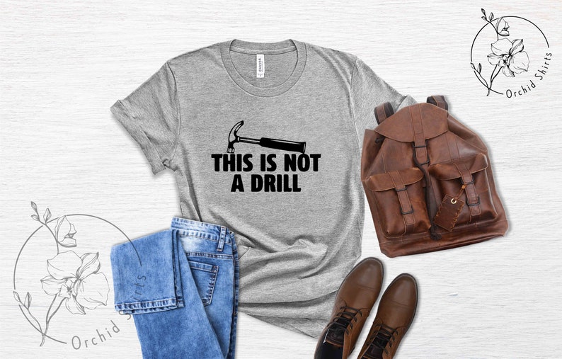 May include: A gray t-shirt with a black hammer and the text "This is not a drill". The shirt is laid out on a wooden surface with a pair of jeans, a brown leather backpack, and brown leather boots.