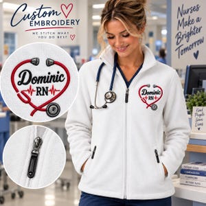 May include: A white fleece zip-up jacket featuring an embroidered red stethoscope shaped like a heart with the name Dominic and the letters RN in black text. The jacket includes a high collar, long sleeves, and two zippered side pockets.