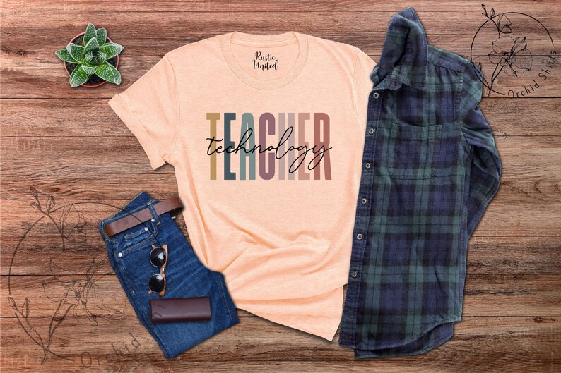 Pu&ograve; includere: Una maglietta color pesca con il testo "TEACHER technology" in strisce verticali colorate. La maglietta &egrave; appoggiata su una superficie di legno con un paio di jeans blu, una cintura marrone, occhiali da sole e una custodia marrone.