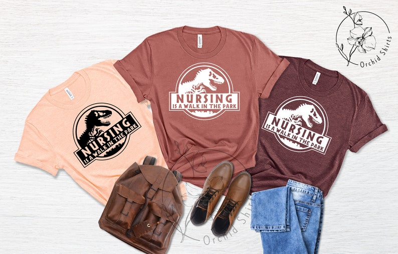 May include: Three t-shirts with a dinosaur graphic and the text "Nursing is a walk in the park". The shirts are in peach, maroon, and burgundy.