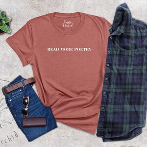 Op de afbeelding: Een roestkleurig T-shirt met de tekst "READ MORE POETRY" in witte letters. Het T-shirt ligt op een lichtgrijs, getextureerd oppervlak met een blauwe jeans, een bruine leren riem, een zonnebril en een bruine leren etui.