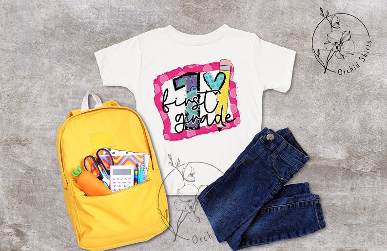 May include: A white t-shirt with a pink frame and the text "1st grade" inside. The number 1 is decorated with colourful shapes and a pencil. A yellow backpack with school supplies is in the background.