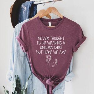 May include: A maroon t-shirt with the text "Never thought I'd be wearing a unicorn shirt but here we are" and a white outline of a unicorn.