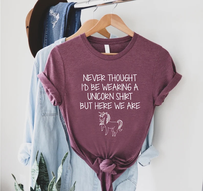 May include: A maroon t-shirt with the text "Never thought I'd be wearing a unicorn shirt but here we are" and a white outline of a unicorn.