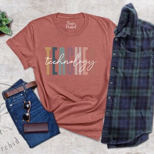 Pu&ograve; includere: Una maglietta color ruggine con il testo "TEACH" in colori arcobaleno e la parola "tecnologia" in bianco sotto. La maglietta &egrave; appoggiata su una superficie grigia testurizzata con un paio di jeans blu, una cintura di cuoio marrone e un paio di occhiali da sole.