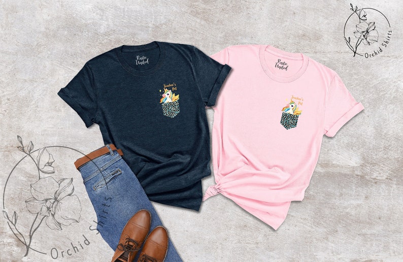 May include: Two t-shirts, one navy blue and one pink, with a pocket design featuring a unicorn and the text "Teacher's Pet." The pocket has a leopard print pattern. The shirts are displayed with jeans and brown shoes.
