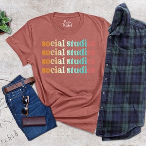 May include: A terracotta-colored t-shirt with the words "social studies" stacked in a retro font. The shirt is paired with blue jeans, a brown belt, sunglasses, and a wallet. A plaid shirt and a small potted plant complete the ensemble.