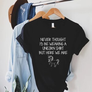 May include: Black t-shirt with white text that reads "Never thought I'd be wearing a unicorn shirt but here we are." A white outline of a unicorn is below the text.